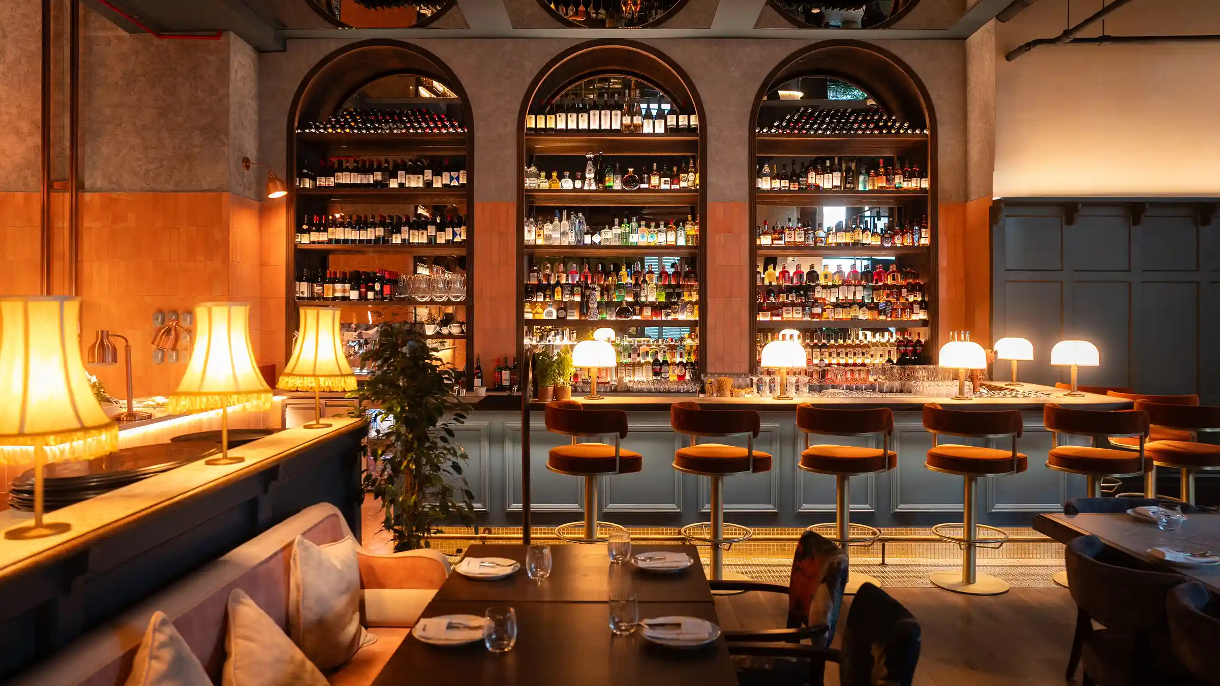 A warm and stylish bar-restaurant space with classic lighting, plush seating, and a vibrant display of fine spirits.